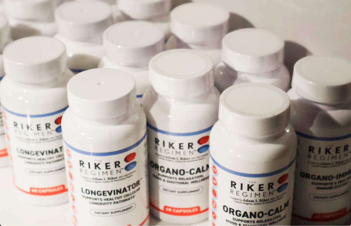 A close-up, slightly angled view of several white plastic supplement bottles from the brand Riker Regimen, including Longevinator and Organo-Calm, arranged in rows on a light surface.