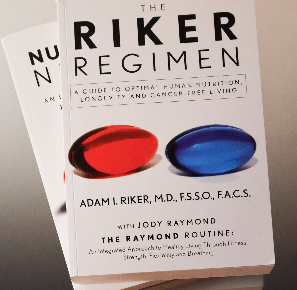 Copies of the books 'THE RIKER REGIMEN' and 'NUTRITIONAL NIRVANA' stacked on a light surface, with the front copy showing a white cover, large black title text, and two pill illustrations in red and blue.